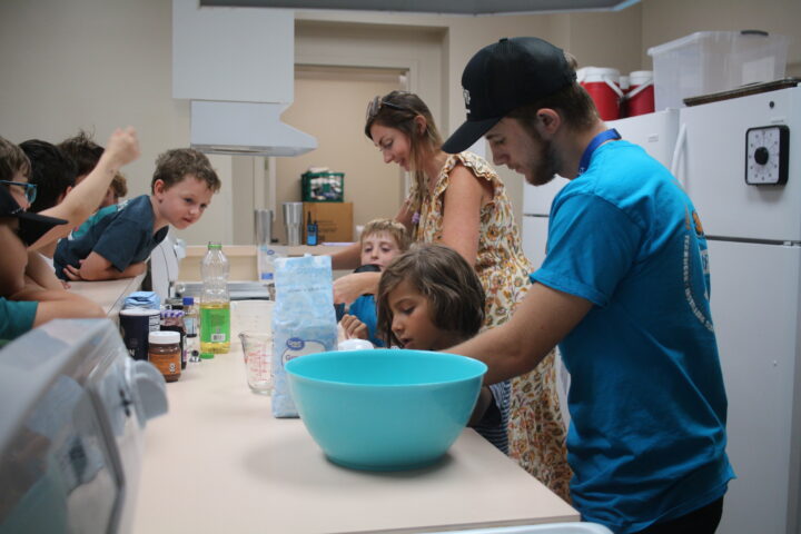 A group of children and two adults are gathered around a kitchen counter with baking ingredients and a large blue bowl. One adult is assisting a child in the mixing process.