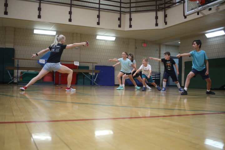 Instructor leads a group of children in a lunge exercise in a gym.