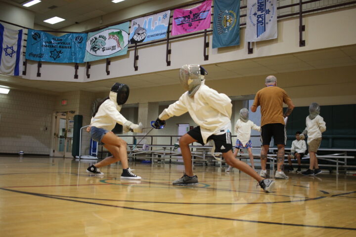 Two young people in fencing gear face off in a gymnasium. Others practice in the background while a coach observes. Colorful banners hang from a balcony above.