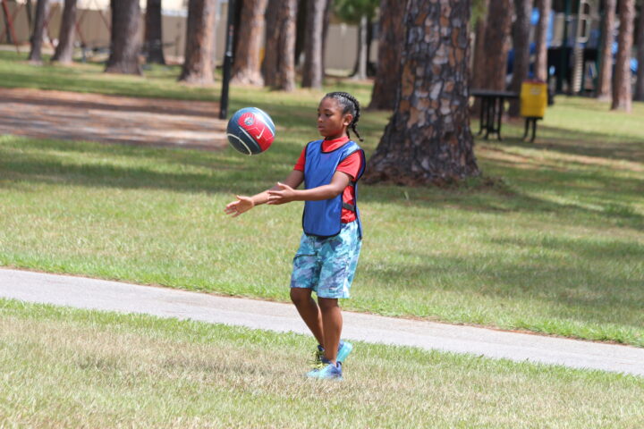 A child in a blue jersey and colorful shorts plays catch with a red and blue ball in a park with trees and grass.