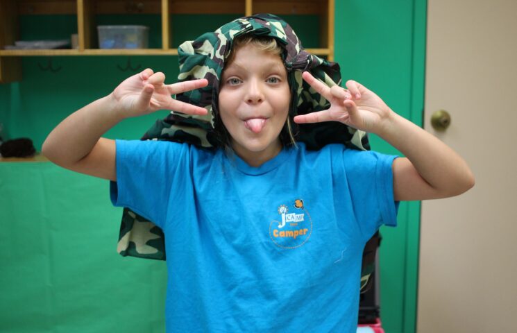 Child in a blue t-shirt making a playful peace sign gesture with both hands, tongue out, and wearing a camouflage fabric on their head.