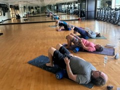 People lying on yoga mats in a gym, using foam rollers under their backs, with exercise bikes visible in the background.