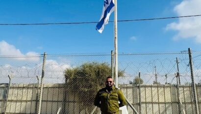 A person in military uniform stands in front of a flagpole with an Israeli flag, against a backdrop of wire fencing and a clear blue sky.