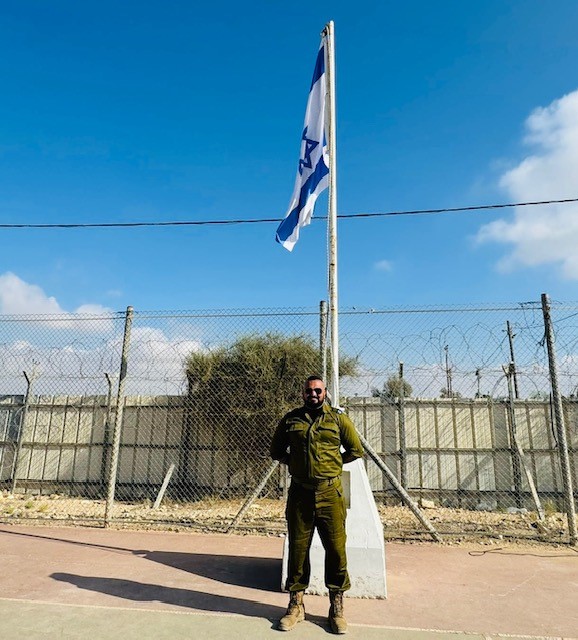 A person in military uniform stands in front of a flagpole with an Israeli flag, against a backdrop of wire fencing and a clear blue sky.