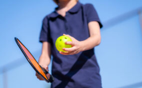 A person in a navy shirt holds a paddle and a yellow perforated ball outdoors.