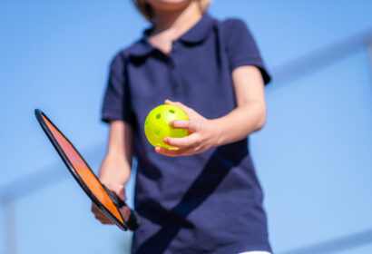 A person in a navy shirt holds a paddle and a yellow perforated ball outdoors.