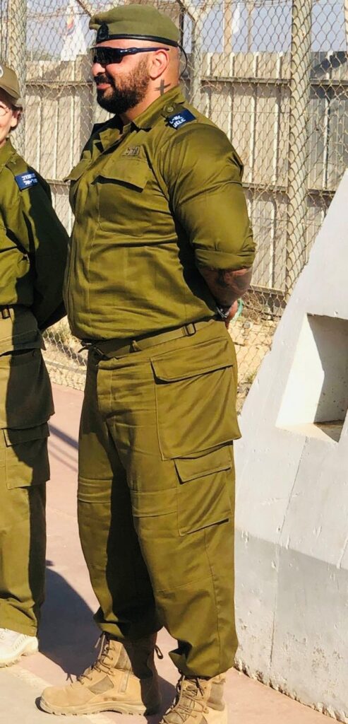 Person in olive green military uniform with patches, wearing sunglasses and a cap, standing outdoors near a fence.