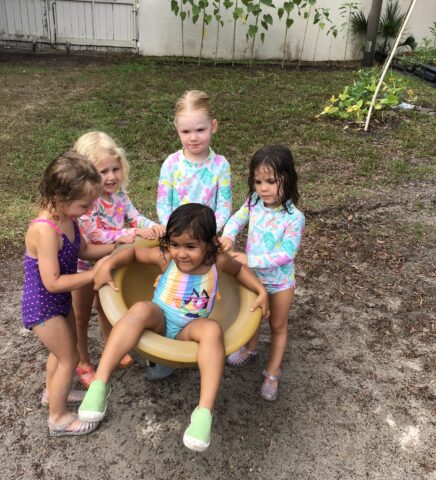 Five children play outside, with four standing around a seated girl in a small yellow bowl. They are all wearing swimsuits and appear to be enjoying a playful moment.