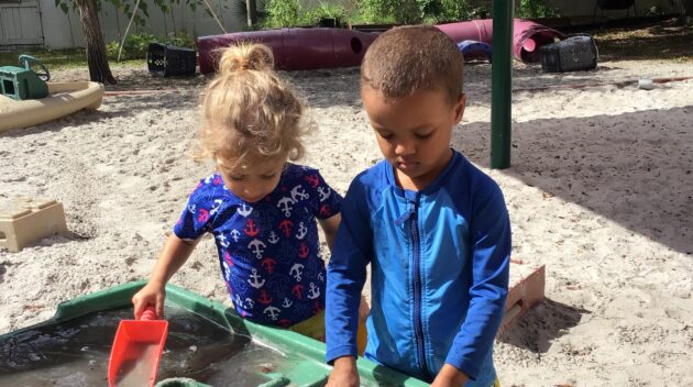 Two children playing with water at a sand table outdoors, surrounded by trees and playground equipment.