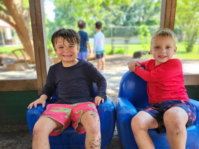 Two children sit in blue chairs outdoors, wearing wet and muddy clothes, smiling. Other children play in the background.