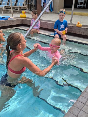 A woman assists a child in pink goggles swimming in a pool. Another child sits on the edge with a yellow toy.