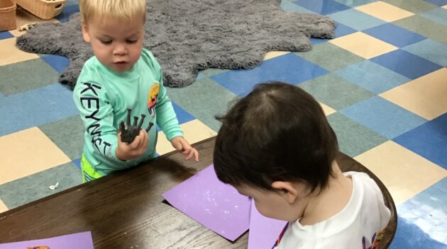 Two toddlers at a table; one holds a toy animal while the other looks down. Purple paper is on the table. Classroom setting with tiled floor and rug.