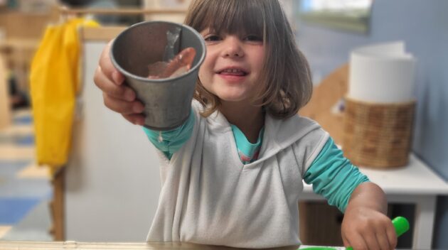 Child holding a metal cup and a net, smiling while standing at a water play table in a classroom setting.