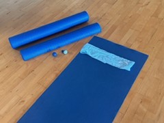 A blue yoga mat, two foam rollers, two massage balls, and a folded towel are arranged on a wooden floor.