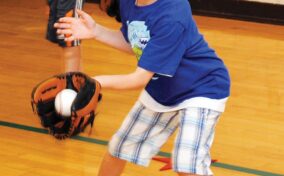 Child in blue shirt and plaid shorts holds a baseball in a mitt, standing on a gym floor.