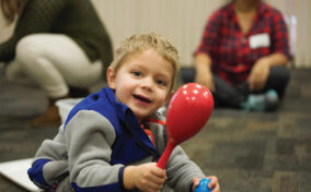 A young child in a blue and gray jacket holds a red maraca and smiles. Two adults sit blurred in the background on a carpeted floor.