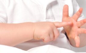 A baby wearing a white shirt holds up one hand and points at it with the other hand.