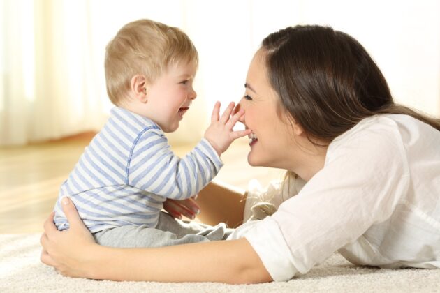 A baby in a striped shirt touches a smiling woman's nose while they both lie on a carpeted floor in a brightly lit room.