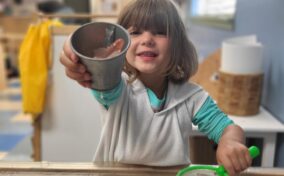 A child smiling, holding a metal cup and a green net over a water play table indoors.