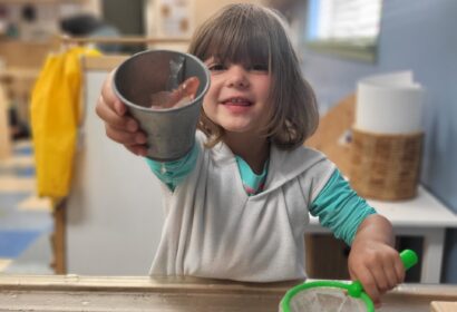 A child smiling, holding a metal cup and a green net over a water play table indoors.