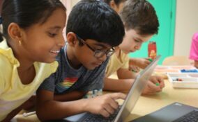 Three children are gathered around a laptop, focused and engaged, with various classroom supplies on the table.