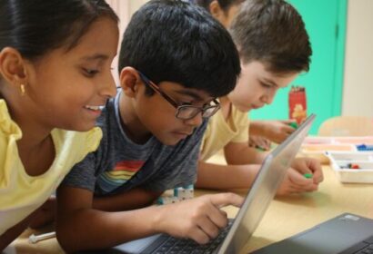 Three children are gathered around a laptop, focused and engaged, with various classroom supplies on the table.