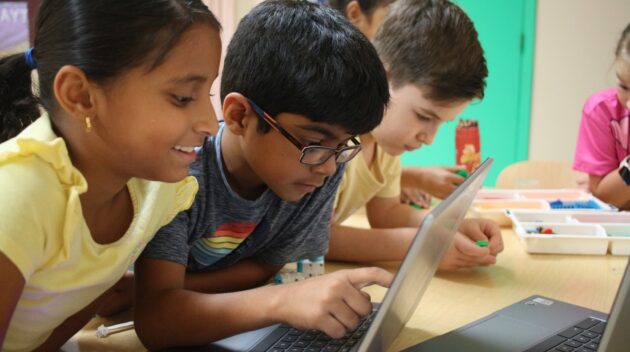 Three children are gathered around a laptop, focused and engaged, with various classroom supplies on the table.