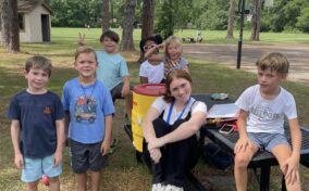 A group of children and one adult sitting and standing around a picnic table outdoors in a park. Trees and a basketball hoop are in the background.