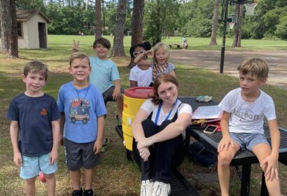 A group of children and one adult sitting and standing around a picnic table outdoors in a park. Trees and a basketball hoop are in the background.