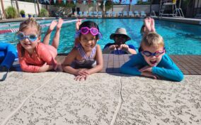 Four children in swimwear and goggles pose at the edge of a swimming pool, with an adult in a hat partially submerged in the background.