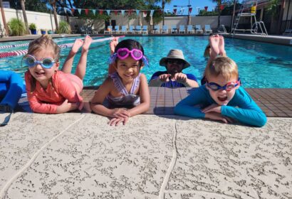 Four children in swimwear and goggles pose at the edge of a swimming pool, with an adult in a hat partially submerged in the background.