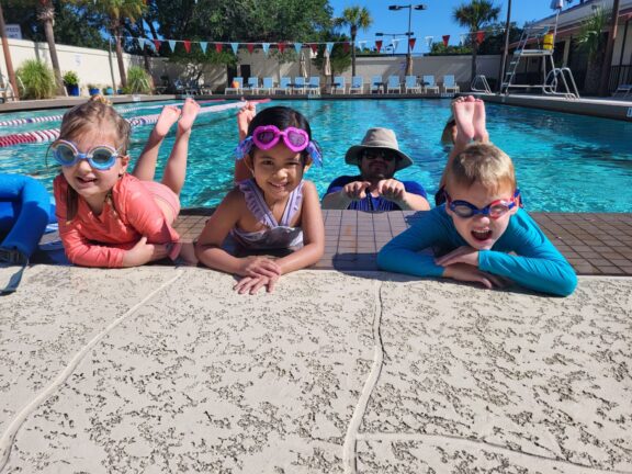 Four children in swimwear and goggles pose at the edge of a swimming pool, with an adult in a hat partially submerged in the background.