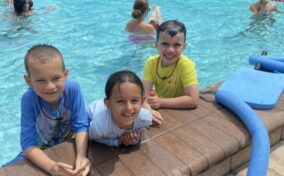 Three children are smiling at the edge of a swimming pool. More people are swimming in the background. Pool toys are nearby.