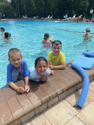 Three children are smiling at the edge of a swimming pool. More people are swimming in the background. Pool toys are nearby.