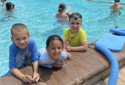 Three children are smiling at the edge of a swimming pool. More people are swimming in the background. Pool toys are nearby.