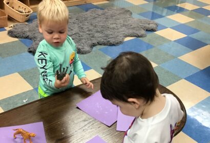 Two children at a table, one is holding a toy animal, and the other is looking down at purple paper.