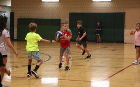 Children playing basketball in a gymnasium, with one boy holding a ball and others moving around the court.