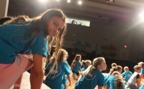 Children in blue shirts gather on a stage with bright overhead lights. A girl in the foreground looks toward the camera.