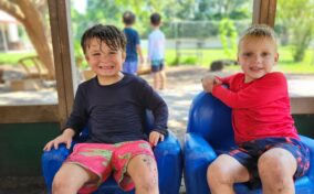 Two children sit smiling on blue chairs outside, wearing swimsuits. The child on the left has wet hair. Two more children are visible in the background. The scene is sunny and vibrant.