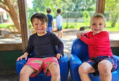 Two children sit smiling on blue chairs outside, wearing swimsuits. The child on the left has wet hair. Two more children are visible in the background. The scene is sunny and vibrant.