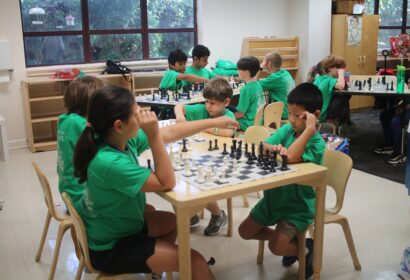 Children wearing green shirts are playing chess on wooden tables in a classroom setting. There are multiple games happening simultaneously, with focused expressions on the players' faces.