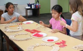 Three children sit at a table painting clothespins red, with circular arrangements of clothespins on the table and red paint on plates. Supplies and doors are visible in the background.