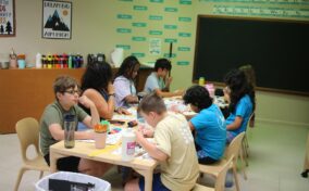 A group of students seated around a table in a classroom, engaged in art activities with various supplies.