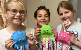 Three children smiling and holding colorful jellyfish crafts with ribbon tentacles in a classroom setting.