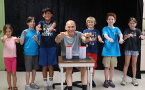 A group of children and a bald man with paint on their hands pose behind a table with two paint cans in front of a black backdrop.