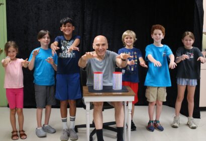 A group of children and a bald man with paint on their hands pose behind a table with two paint cans in front of a black backdrop.