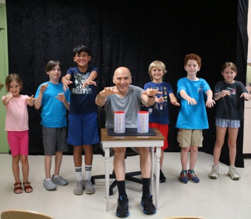 A group of children and a bald man with paint on their hands pose behind a table with two paint cans in front of a black backdrop.