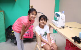 Two girls in a room; one smiles while standing, the other sits near a sewing machine on a table. Box storage and a green wall are in the background.