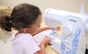 A child operates a sewing machine to stitch a colorful fabric square in a classroom setting.