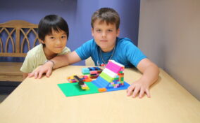 Two children sitting at a table, smiling, with a colorful Lego structure in front of them.